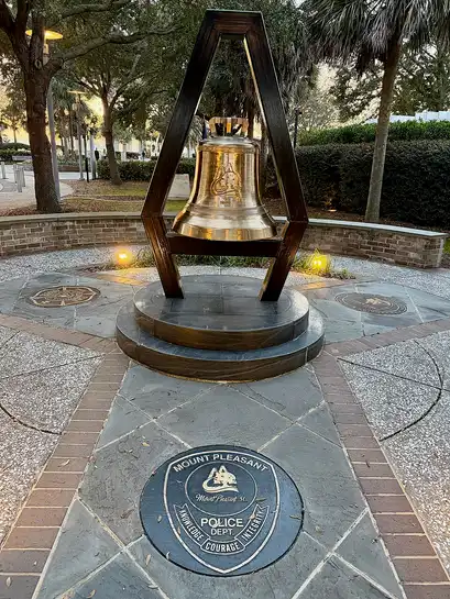 A bronze bell hung in a tall frame above a paved circle with an inlaid emblem, at a waterfront park