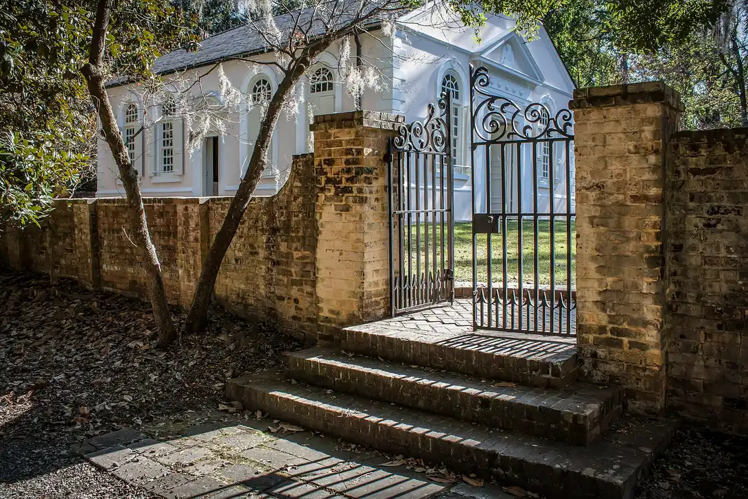 The white stucco St. James Goose Creek church seen through an open iron gate in a brick wall