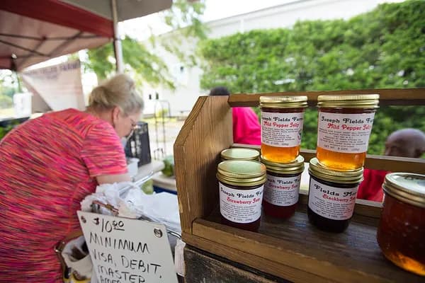 A vendor arranging jars of local honey on a wooden stand under a tent at the North Charleston Farmers Market
