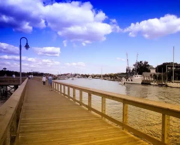 A wooden boardwalk with lampposts running beside Shem Creek, with shrimp boats and waterfront buildings beyond