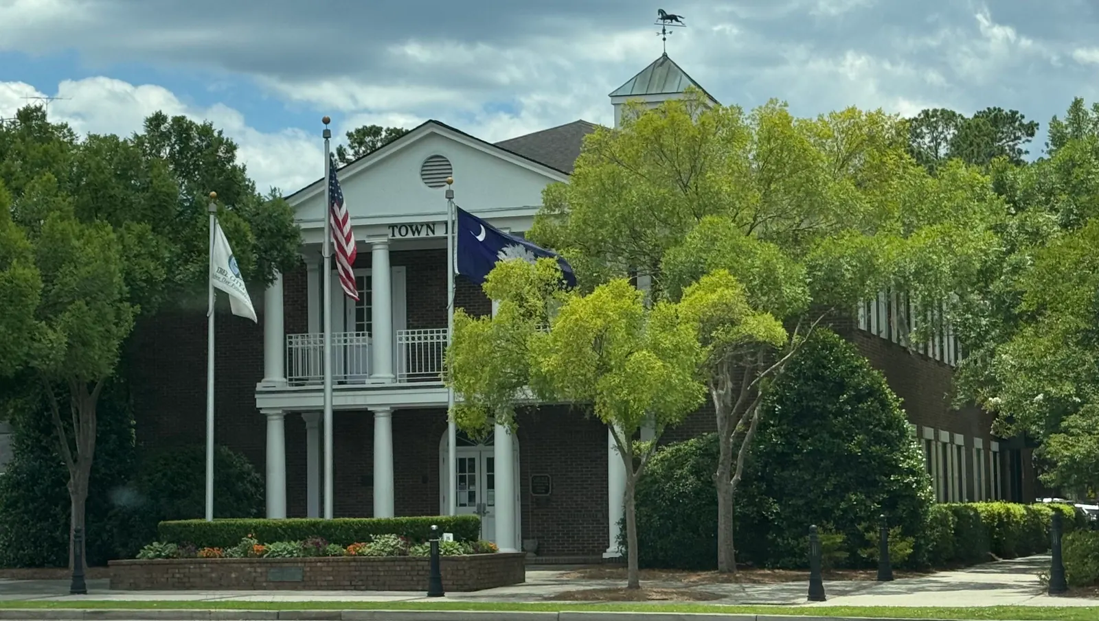 White columned town hall building with a cupola and flags, framed by trees, in Summerville