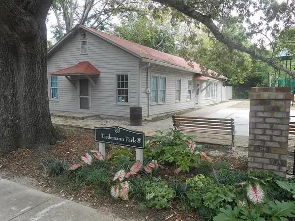 A small gray wooden building with a red awning behind a garden bed and the Tiedemann Park sign