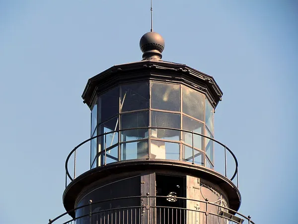 The black lantern room and gallery at the top of the Hunting Island Lighthouse against a pale blue sky