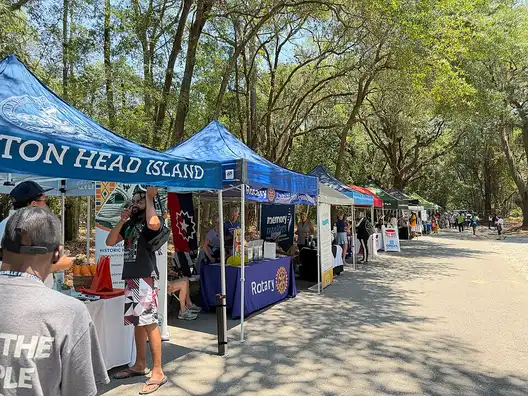 Blue vendor tents lined up under live oaks along a shell path at Historic Mitchelville Freedom Park during a Juneteenth celebration