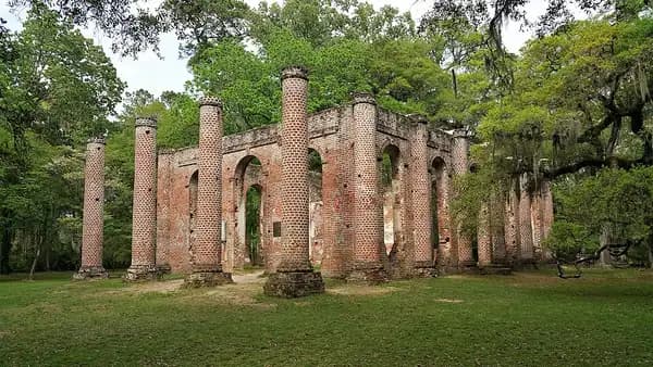 Brick columns and arched walls of the roofless Old Sheldon Church ruins standing in a green clearing among oaks