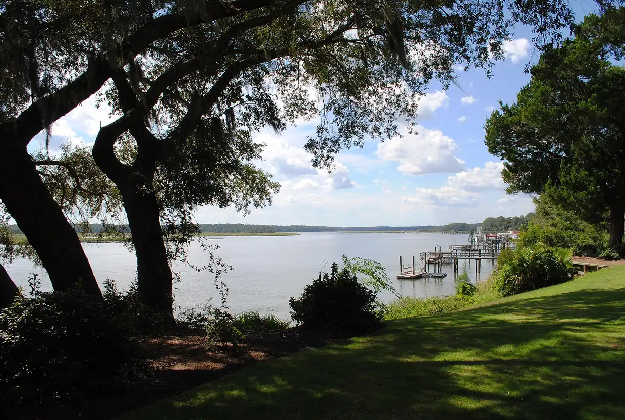 The May River seen past live oak branches from a grassy bluff, with docks and marsh along the far shore