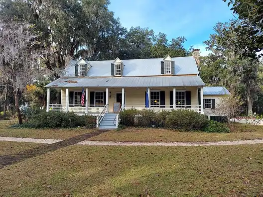 The white Heyward House with a front porch and metal roof, set back on a lawn under trees in Old Town Bluffton